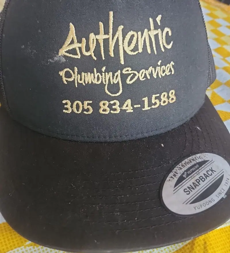 Black cap embroidered with the gold Authentic Plumbing Services logo and phone number