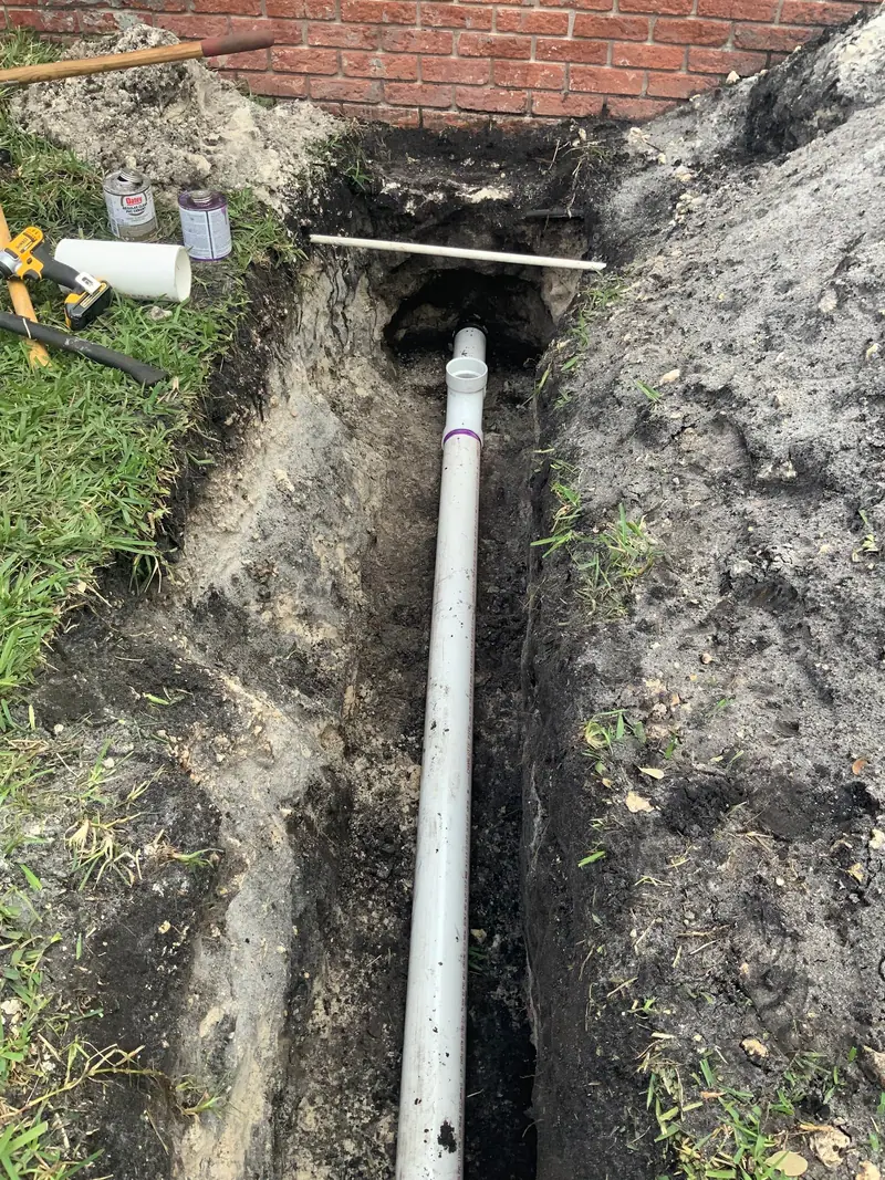 Long trench beside a brick house with a new white PVC line next to the old black pipe